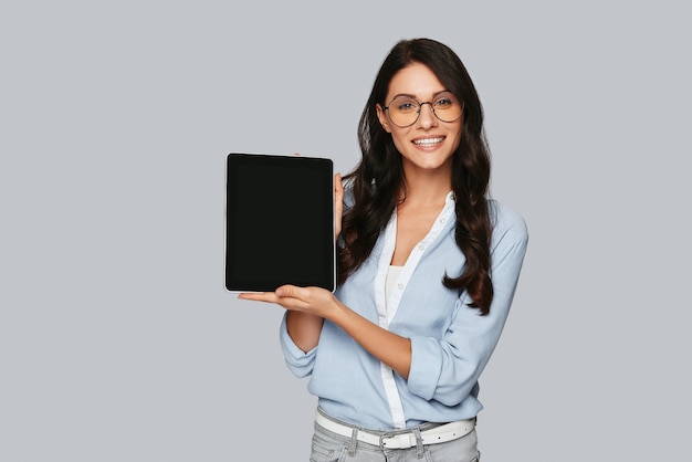 Guarda qui! Attraente giovane donna che punta lo spazio della copia sulla tavoletta digitale e sorride mentre si trova in piedi su sfondo grigio gray
