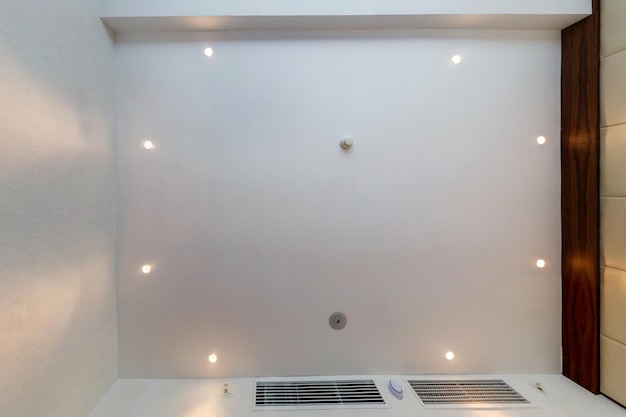 Guarda su controsoffitto con lampade alogene e costruzione in cartongesso con sensore di allarme antincendio in una stanza vuota in un appartamento o in una casa Soffitto teso bianco e di forma complessa