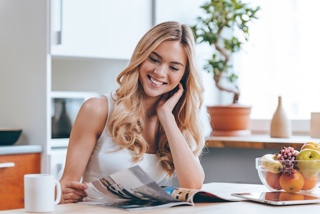 Guardando attraverso la rivista al mattino. Bella giovane donna allegra che legge una rivista mentre è seduta in cucina a casa