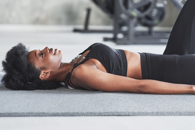 Guardando in alto quando si sdraia sul pavimento e si prende una pausa, la donna afroamericana con i capelli ricci e in abiti sportivi ha una giornata di fitness in palestra