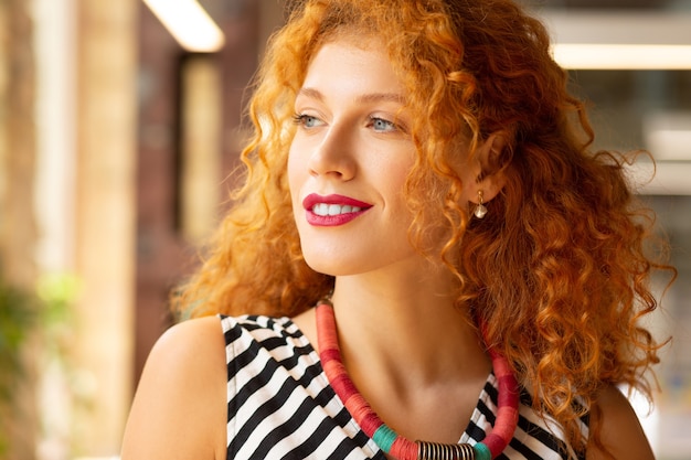 Guardando nella finestra. Donna riccia dai capelli rossi che indossa una collana guardando nella finestra