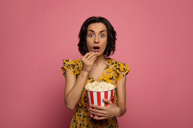 Guardare dei film. giovane donna sorpresa, che sta mangiando popcorn e guardando nella telecamera.