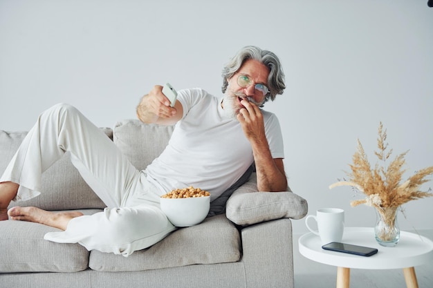 Guardare programmi TV e mangiare popcorn Uomo moderno ed elegante con capelli grigi e barba al chiuso