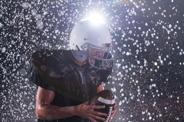 Guerriero atleta di football americano in piedi su un campo tiene il suo casco e pronto a giocare. Giocatore che si prepara a correre, attaccare e segnare touchdown. Notte piovosa con riflessi sull'obiettivo drammatici e gocce di pioggia. Alto