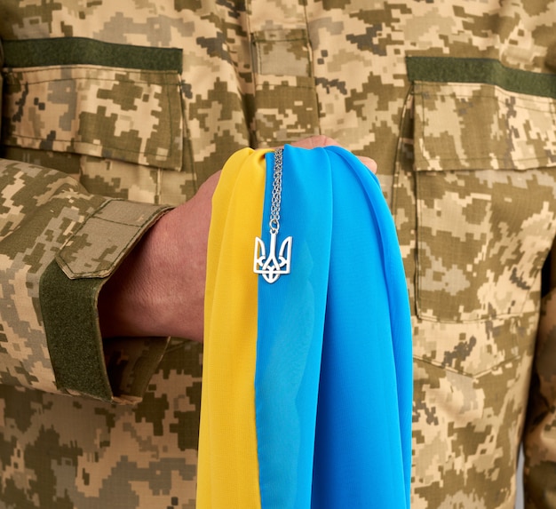 Guerriero ucraino vestito con un'uniforme militare a pixel detiene la bandiera giallo-blu dello stato dell'Ucraina e sulla catena un piccolo stemma del paese di tridente