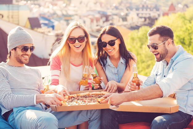 Gustare la pizza con gli amici. Quattro giovani allegri che mangiano pizza e bevono birra seduti ai sacchi di fagioli sul tetto dell'edificio