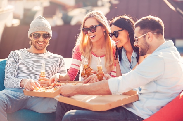 Gustare la pizza insieme. Quattro giovani allegri che mangiano pizza e bevono birra seduti ai sacchi di fagioli sul tetto dell'edificio