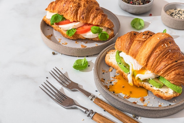 Gustosa colazione con panini croissant con uovo in camicia avocado formaggio morbido mozzarella e pomodoro in un piatto con forchette su sfondo bianco