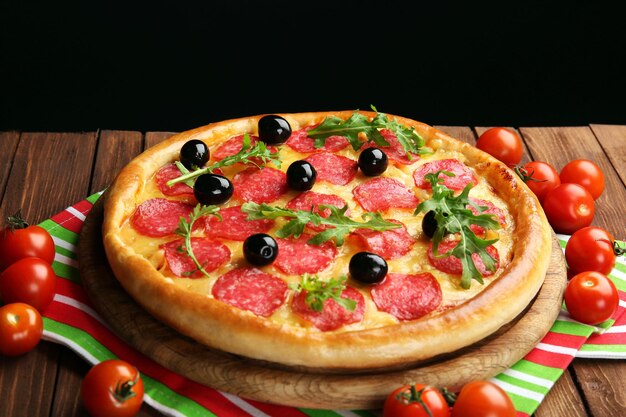 Gustosa pizza con salame e olive decorate con pomodori su tavola di legno su sfondo nero