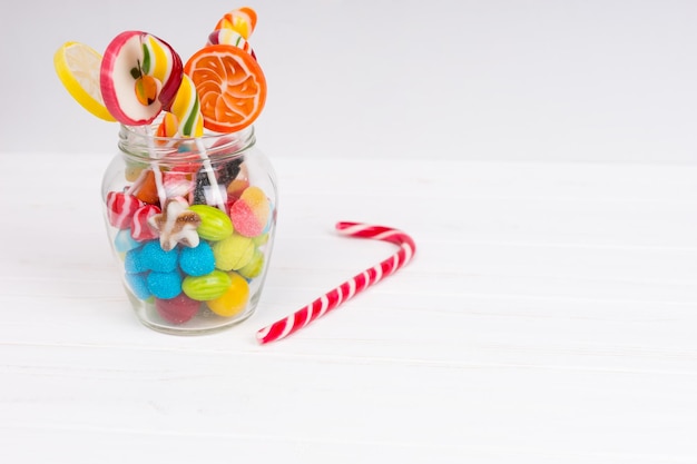 Gustosi dolci da masticare, lecca-lecca brillanti e caramelle di gelatina nel bicchiere vicino al bastoncino di zucchero su una tavola bianca di legno