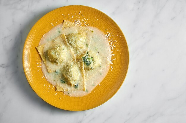 Gustosi ravioli cotti con vari ripieni in salsa cremosa cosparsa di parmigiano su piatto giallo brillante su marmo bianco