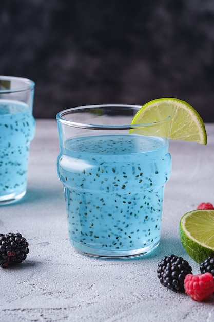 Gustoso cocktail di colore blu con semi di chia basilico, fetta di limone e agrumi, bacche di lampone e mora in due bicchieri, bevanda estiva sana, tavolo in pietra