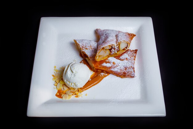 Gustoso dessert dolce. Strudel di mele arioso con zucchero a velo, marmellata di cioccolato, fette sottili di nocciole e una porzione di gelato alla panna bianca. Sfondo nero