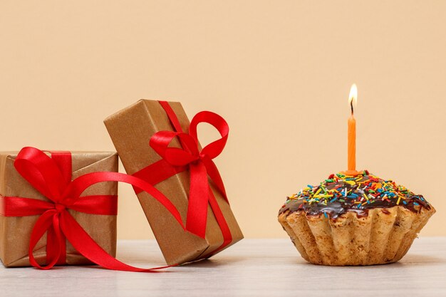 Gustoso muffin di compleanno con glassa al cioccolato e caramello, decorato con candele festive accese e scatole regalo su fondo beige. Buon compleanno concetto minimo.