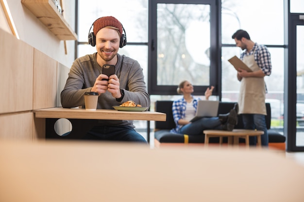 Gustoso spuntino. Piacevole bell'uomo positivo seduto al tavolino del bar e usando il suo smartphone mentre mangia uno spuntino gustoso