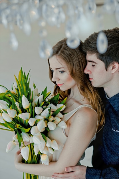 Guy abbraccia la ragazza da dietro. ragazza con bouquet di tulipani in mano da ragazzo a San Valentino. la coppia innamorata si trova sotto un bellissimo lampadario.