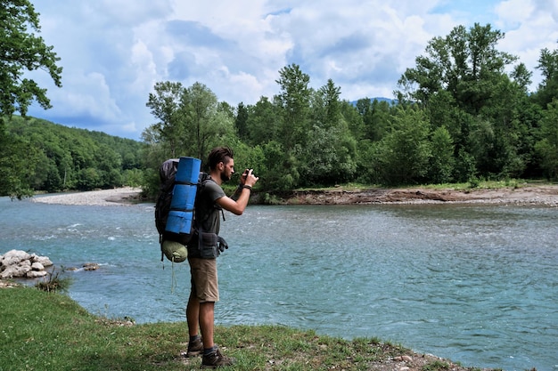 Guy è un viaggiatore si trova sulla riva del fiume di montagna con un grande zaino da trekking e scatta foto