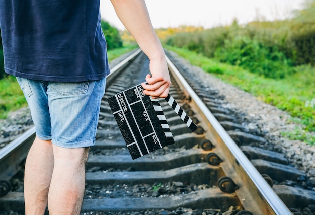 Guy tiene in mano un ciak nero. L'uomo sta dirigendo e filmando un film amatoriale. Sentieri ferroviari sullo sfondo, concetto di viaggio.