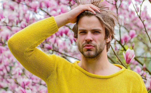 Ha mal di testa dall'odore uomo con la barba lunga albero di magnolia ragazzo sexy fiore in fiore albero primavera stile moda Sessualità maschile sakura sfondo godersi il bel tempo fiore fresco fiore