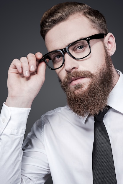 Ha una mente creativa. Bel giovane uomo barbuto in camicia e cravatta che si aggiusta gli occhiali e guarda la telecamera mentre sta in piedi su sfondo grigio