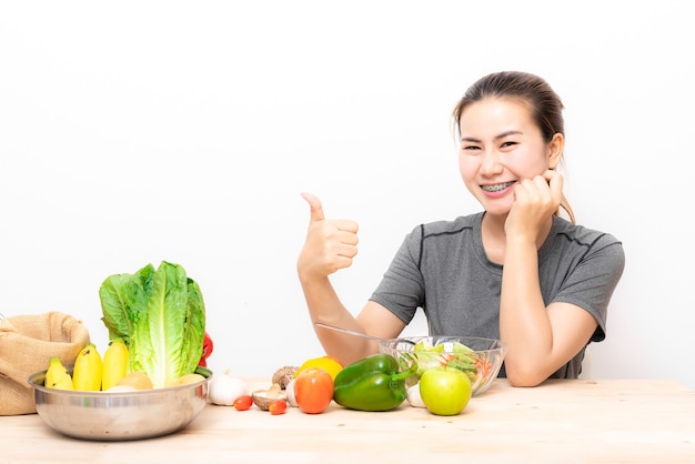 Haapy donna asiatica con parentesi graffe dentali che mostra il pollice in su mentre si mangia verdura insalata su bianco