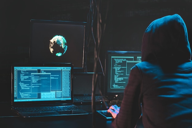 Hacker incappucciato che utilizza software dannoso hackera il data center aziendale malfattore nascosto sottoterra in un luogo oscuro display multipli con codice di phishing e attacco globale alle mappe