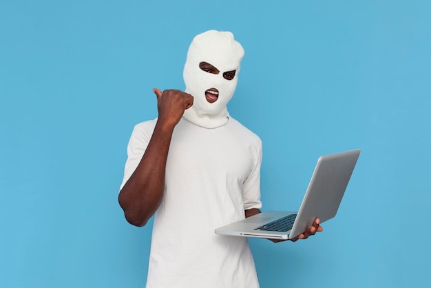 hacker maschio afroamericano in balaclava bianca usa il portatile e indica sullo sfondo blu