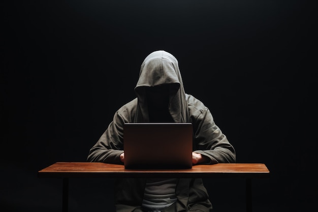 Hacker senza volto in una cappa con un laptop su uno sfondo nero