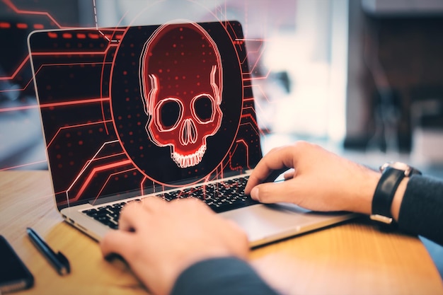 Hacking attack and online piracy creative concept with red skull digital hologram on modern laptop screen background and male hands on a keyboard double exposure