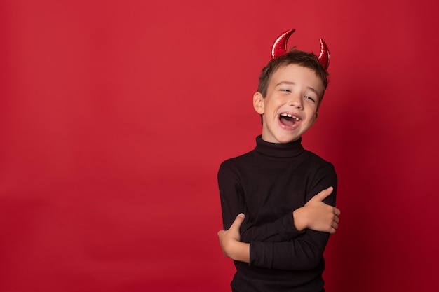 Halloween Il bambino felice ride ad alta voce con corna diaboliche su uno sfondo rosso da studio