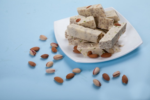 Halva a base di semi di girasole, mandorle e pistacchi giace su un piatto su uno sfondo blu. Pistacchi e mandorle sono sparsi uno accanto all'altro.