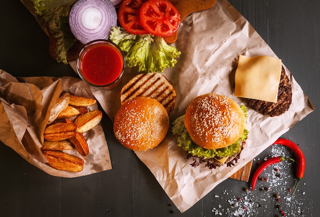 Hamburger casalingo fresco delizioso su una tavola di legno. accanto al componente per hamburger, vassoi di legno, patate fritte e peperoncino. Un bicchiere di succo di pomodoro