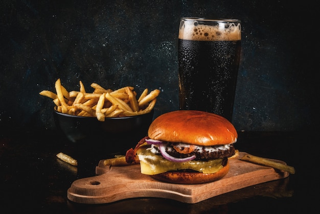 Hamburger del formaggio e della carne fresca con le patate fritte e il vetro della birra scura dello zenzero, su blu scuro, copyspace