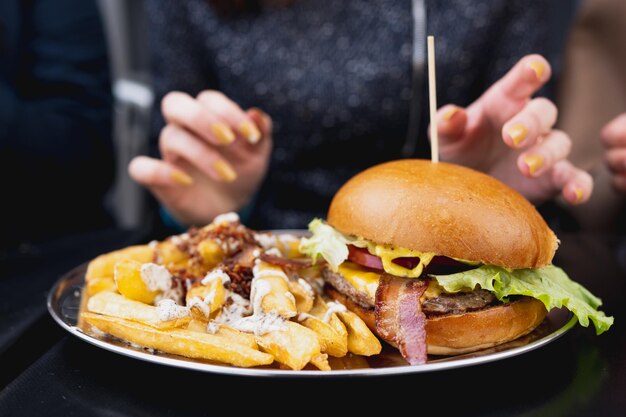 Hamburger di bacon gourmet di primo piano con patatine fritte sul tavolo e mani femminili irriconoscibili sullo sfondo.