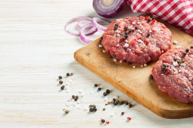 Hamburger di bistecca di manzo tritato fresco crudo fatto in casa con spezie e cipolla su un tagliere, su un tavolo di legno bianco