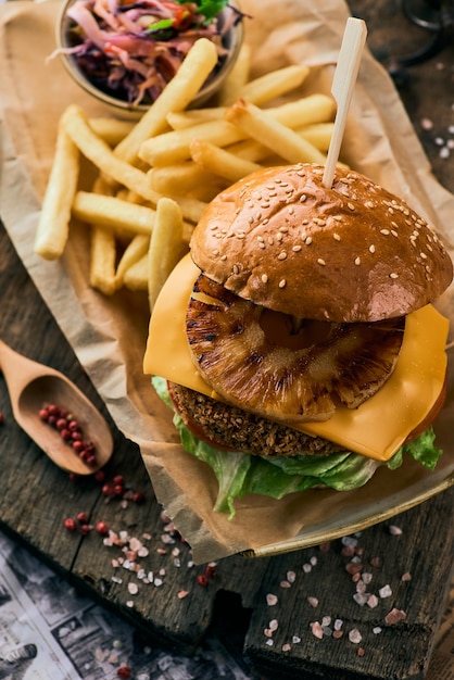 Hamburger di sesamo con ananas, formaggio e patatine fritte su un tagliere di legno.