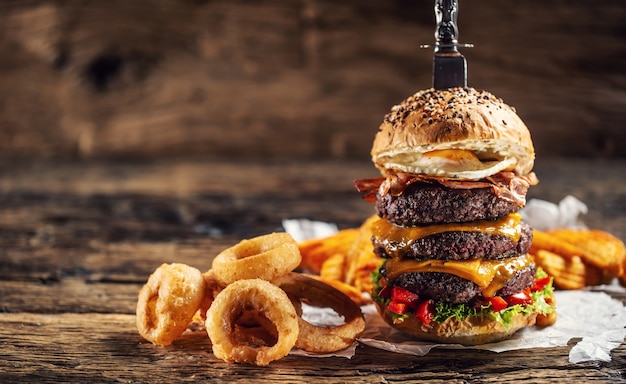 Hamburger di triplo manzo con formaggio fuso, uova e anelli di cipolla su legno rustico.