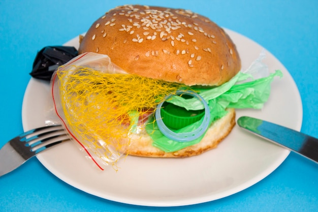 Hamburger fresco e gustoso con rifiuti di plastica all'interno su piatto bianco su sfondo blu