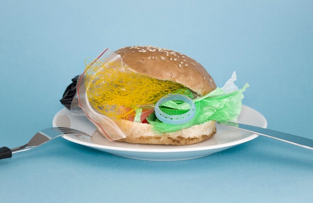 Hamburger fresco e gustoso con rifiuti di plastica all'interno su piatto bianco su sfondo blu