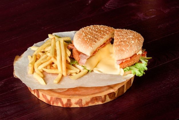 Hamburger in un taglio con cotoletta, formaggio fuso, pancetta, lattuga, pomodori e patate su pergamena su un piatto di legno su un tavolo di legno.