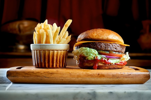 Hamburger succoso e appetitoso con pomodori, lattuga, cetriolo, formaggio fuso e cipolle con pan brioche servito su una tavola di legno con patatine fritte