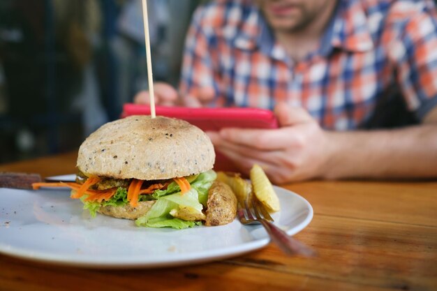 Hamburger vegetariano di patate dolci con verdure fresche servito su piatto bianco Uomo caucasico sfocato seduto sullo sfondo