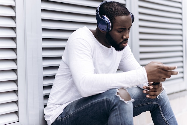 Handsome afro americano che indossa abiti casual nella città moderna ascoltando musica con le cuffie