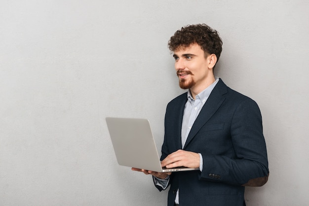 Handsome giovane imprenditore fiducioso che indossa una giacca in piedi isolato su grigio, lavorando su un computer portatile