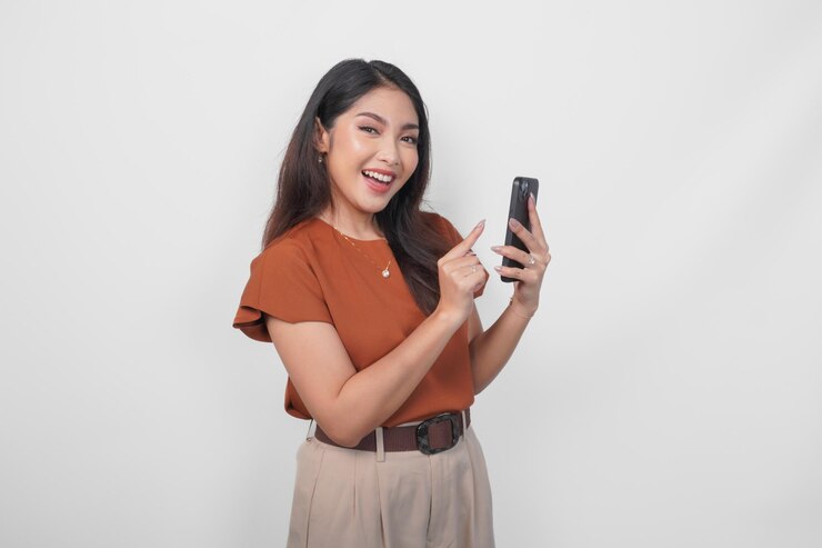 Happy Asian woman is smiling and pointing to her smartphone wearing brown shirt isolated by a white background