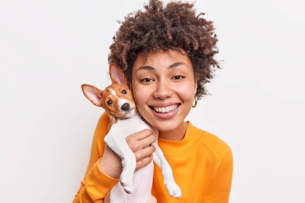 Happy dark skinned young woman expresses care and responsibility to her favorite small puppy gets pleasure while playing with dog smiles broadly have fun together isolated over white wall