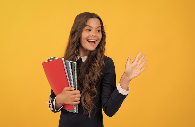 Happy teen girl tenere il quaderno scolastico per i compiti che studiano su sfondo giallo salutando la scuola