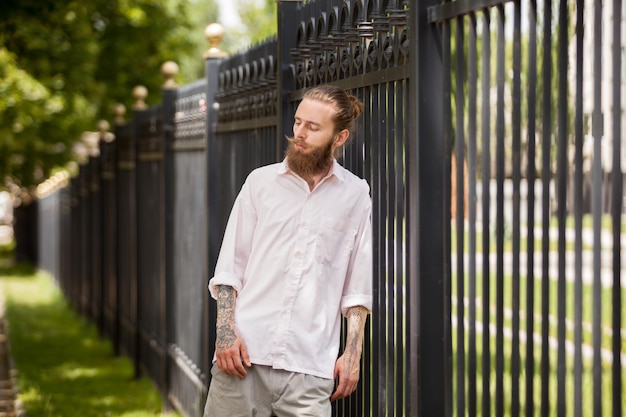 Hipster con barba lunga in posa accanto a una recinzione fuori città. Stile e diversità