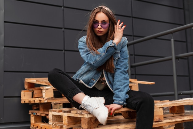 Hipster di giovane donna alla moda in occhiali viola giovanili in giacca di jeans blu alla moda in jeans neri in stivali di pelle bianca sta riposando su pallet di legno in città. Ragazza moderna sexy all'aperto.
