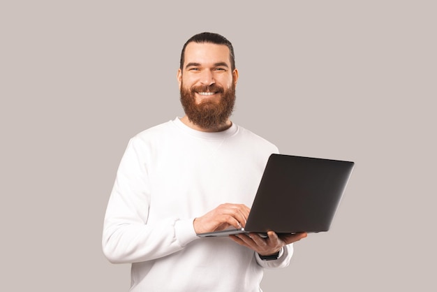 Hipster maschio barbuto tiene e digita su un laptop mentre sorride alla telecamera Studio girato su sfondo grigio